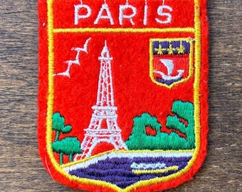 Può includere: Una toppa ricamata rettangolare con sfondo rosso e bordo giallo. Presenta la scritta bianca PARIS in alto, una torre Eiffel bianca, alberi verdi, un fiume blu e un piccolo stemma con una barca bianca e tre stelle gialle.