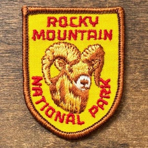 May include: Embroidered patch featuring a ram's head and the text "Rocky Mountain National Park." The patch has a yellow background with red and brown text and border. The ram is brown and tan.