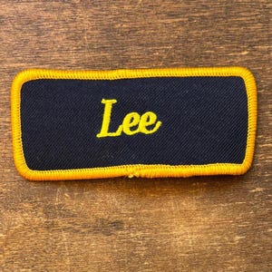 May include: A rectangular patch with the name "Lee" embroidered in yellow script on a navy blue background. The patch has a yellow border and is set against a wooden surface.