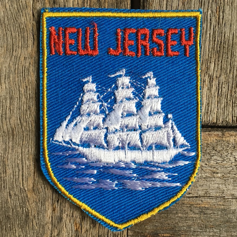 May include: A shield-shaped embroidered patch with a blue background and a yellow border. The text NEW JERSEY is stitched in red at the top. Below the text is a white three-masted sailing ship on stylised white waves, all rendered in thread.