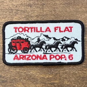 Può includere: Toppa rettangolare con bordo nero, con il testo "TORTILLA FLAT" e "ARIZONA POP. 6" in rosso. Il disegno include una diligenza rossa trainata da quattro cavalli neri, sullo sfondo di montagne.