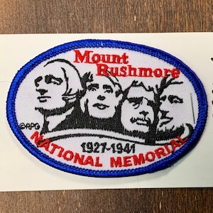 May include: A white oval patch with a blue border. The patch features a black and white image of the Mount Rushmore National Memorial, with the text "Mount Rushmore" in red and "National Memorial" in red. The text "1927-1941" is also in red.