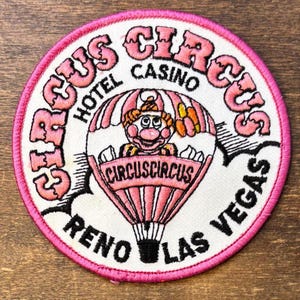 May include: A circular embroidered patch featuring a pink and white hot air balloon with a clown character inside. The text reads CIRCUS CIRCUS HOTEL CASINO RENO LAS VEGAS. The patch has a bright pink stitched border on a white background.
