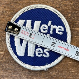 Puede incluir: Un parche bordado circular de color azul con texto blanco bordado que dice We're Wes. El parche presenta un borde blanco rematado y se mide con una cinta m&eacute;trica Craftsman que muestra un di&aacute;metro de aproximadamente 7,6 cm.