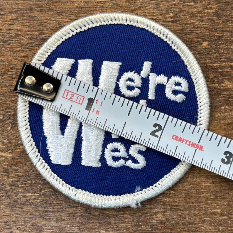 Puede incluir: Un parche bordado circular de color azul con texto blanco bordado que dice We're Wes. El parche presenta un borde blanco rematado y se mide con una cinta m&eacute;trica Craftsman que muestra un di&aacute;metro de aproximadamente 7,6 cm.