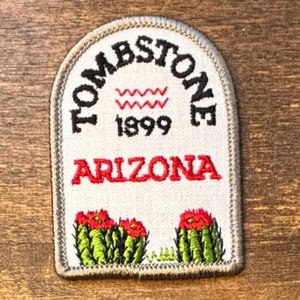 Pode incluir: Um emblema bordado em forma de arco com fundo branco e borda cinza. Apresenta o texto preto "TOMBSTONE", uma linha ondulada vermelha, o ano "1899" em preto, o texto vermelho "ARIZONA" e dois cactos verdes com flores vermelhas.