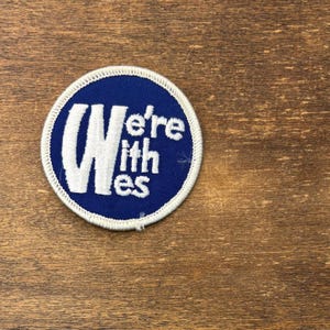 Puede incluir: Un parche bordado circular con un fondo azul intenso y un borde cosido en blanco. El parche presenta el texto blanco "We're with es" dispuesto en un dise&ntilde;o estilizado y apilado en el centro de la superficie circular de tela.