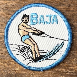 May include: A circular embroidered patch with a white background and a blue stitched border. The design features a person water skiing in blue trunks, holding a tow rope, with the word BAJA embroidered in light blue letters above the figure.