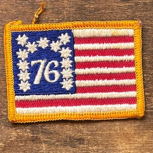 May include: Embroidered patch featuring the American flag design with red and white stripes and a blue field with white stars. The number "76" is embroidered in white. The patch has a gold border and a small loop at the top.