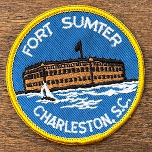 May include: Circular patch with a blue background featuring the text "FORT SUMTER" and "CHARLESTON, S.C." in white. The patch depicts a brown fort with a black flag and white waves.