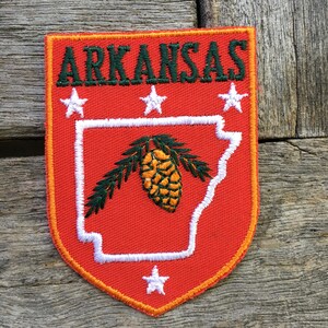 Pode incluir: Um emblema bordado em forma de escudo com fundo laranja e borda amarela. O texto ARKANSAS está bordado em verde escuro no topo. Um contorno branco do estado contém uma pinha e agulhas. Estão incluídas quatro estrelas brancas.