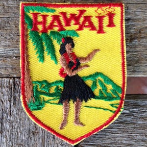 Op de afbeelding: Geborduurde patch met een gele achtergrond en een rode rand. De patch toont het woord "HAWAII" in het rood, een afbeelding van een hula-danseres, palmbomen en bergen. De patch is een souvenir.