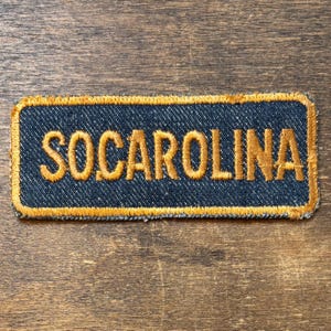 Op de afbeelding: Een rechthoekige patch met het woord "SOCAROLINA" geborduurd in goudkleurig garen op een donkerblauwe denim achtergrond. De patch heeft een gouden rand en is bedoeld om op kleding of accessoires te worden genaaid.