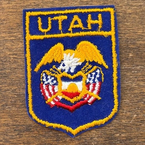 Puede incluir: Un parche azul con la palabra "UTAH" en amarillo en la parte superior. El parche presenta un emblema amarillo y blanco con un diseño en forma de corazón en rojo, blanco y azul. El parche está contorneado en amarillo.