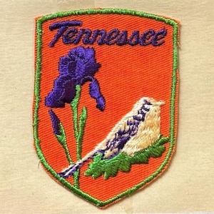 May include: A shield-shaped embroidered patch with an orange background and a green border. It features a purple iris flower with green stems and a cream-colored bird with purple markings perched on green leaves. The word Tennessee is stitched in purple.