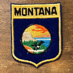 May include: Embroidered Montana patch with a shield shape. The patch features the word "MONTANA" in yellow block letters above a circular design with a mountain, lake, and sunset scene. The patch is bordered with yellow stitching.