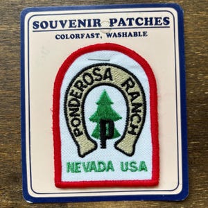 Könnte beinhalten: Ein weißer gestickter Aufnäher mit rotem Rand. Der Aufnäher zeigt ein Hufeisen mit den Worten "Ponderosa Ranch" und einem Kiefernbaum darin. Der Text "Nevada USA" befindet sich unterhalb des Hufeisens.