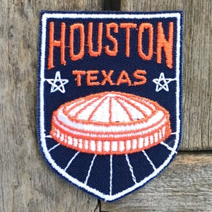 Pode incluir: Um remendo bordado azul e branco com o texto "HOUSTON TEXAS" e uma estrela branca de cada lado. O remendo apresenta uma imagem bordada branca e laranja do Astrodome.