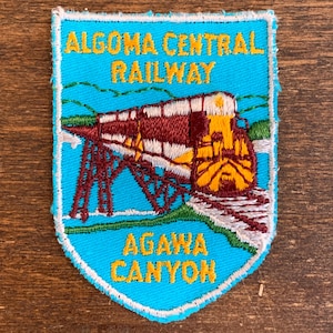 May include: A blue and white embroidered patch with the text "Algoma Central Railway" and "Agawa Canyon". The patch features a red and yellow train on a bridge.