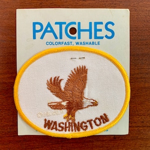 Può includere: Una toppa ovale gialla da stirare con un'aquila ricamata in marrone e il testo "WASHINGTON" in ricamo marrone. La toppa è su una carta bianca con il testo "PATCHES COLORFAST, WASHABLE" in blu.
