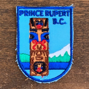 May include: Embroidered patch featuring a totem pole design with the text "PRINCE RUPERT B.C." in blue and white. The patch has a light blue background with a mountain graphic.