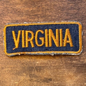 Puede incluir: Un parche rectangular con la palabra "VIRGINIA" bordada en hilo dorado sobre un fondo de mezclilla azul oscuro. El parche tiene un borde dorado, ideal para ropa o accesorios.