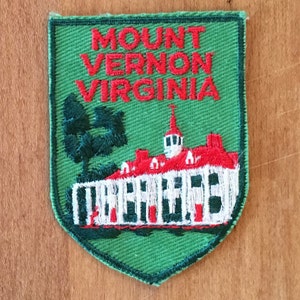 May include: Green embroidered patch with a white building with a red roof and a white steeple. The building is surrounded by a green lawn with a tree. The text "MOUNT VERNON VIRGINIA" is embroidered in red on the patch.