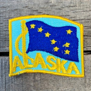 以下が含まれることがあります： アラスカ州旗をあしらった刺繍パッチ。パッチは、8つの金色の星が入った濃い青色の旗と、水色の背景で構成されています。「ALASKA」の文字が旗の下に金色の刺繍で施され、金色の縁取りがされています。