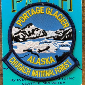 May include: A blue embroidered patch featuring the words "Portage Glacier Alaska Chugach National Forest" in yellow lettering. The patch depicts a mountain range and glacier in white and black.