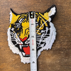 May include: An embroidered tiger head patch featuring yellow, black, white, and red thread. A white measuring tape is placed vertically over the center of the patch, indicating a height of approximately 4 inches from top to bottom.