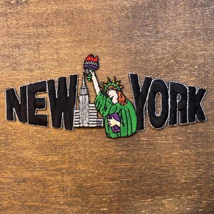 May include: An embroidered patch featuring the text NEW YORK in bold black letters. The letter O is replaced by a white skyscraper and a green Statue of Liberty holding a torch with a red flame and a purple tablet, all outlined in white thread.