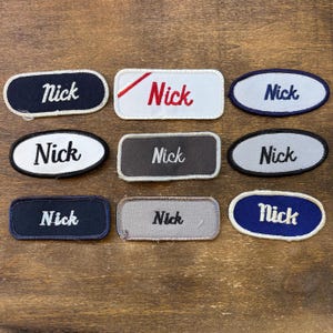 Könnte beinhalten: Neun gestickte Namensschilder mit dem Namen "Nick" in verschiedenen Farben und Stilen. Die Aufnäher sind oval, rechteckig und oval geformt, mit dem Namen in weißem, rotem und blauem Faden gestickt. Sie liegen auf einer Holzoberfläche.