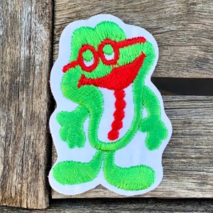 Könnte beinhalten: Gestickter Aufnäher mit einer Cartoon-Froschfigur mit grünem Körper, roter Brille und rotem Schal. Der Aufnäher hat einen weißen Rand und liegt auf einer Holzoberfläche. Der Frosch lächelt.