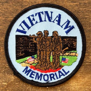 May include: Circular patch with the words "VIETNAM MEMORIAL" in blue. The patch features an illustration of three soldiers in brown uniforms, a brick wall, and two American flags on a green field.