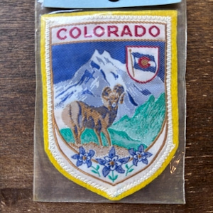 Könnte beinhalten: Ein gestickter Aufnäher in Gelb und Weiß mit dem Wort "COLORADO" in roten Buchstaben. Der Aufnäher zeigt eine Bergkette, ein Dickhornschaf und die Flagge des US-Bundesstaates Colorado.
