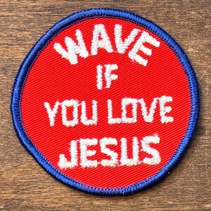 Op de afbeelding: Een ronde patch met een rode kern en een blauwe rand. De patch heeft de witte geborduurde tekst "WAVE IF YOU LOVE JESUS". De patch is waarschijnlijk ontworpen om op kleding of accessoires te worden genaaid.