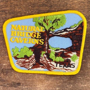 Puede incluir: Parche bordado con una escena de paisaje con el texto "NATURAL BRIDGE CAVERNS TEXAS". El parche tiene un fondo azul con un borde amarillo, que representa un árbol, formaciones rocosas y vegetación.