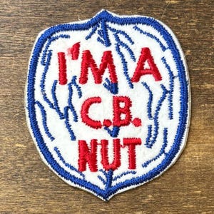 Puede incluir: Un parche bordado en forma de escudo con fondo blanco y borde azul. El texto I'M A C.B. NUT está bordado en hilo rojo en el centro. Un patrón de hilo azul que recuerda a rayos o venas se extiende por toda la superficie blanca.