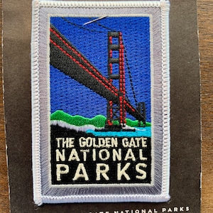 May include: A white embroidered patch with a red and black image of the Golden Gate Bridge. The text "The Golden Gate National Parks" is embroidered in white on a blue background.