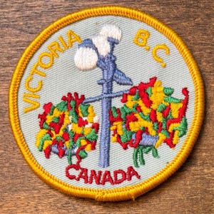 May include: Embroidered patch with a light blue background, featuring a stylized image of a lamp post with colorful floral accents. The patch is circular, with the words "VICTORIA B.C. CANADA" embroidered in gold.