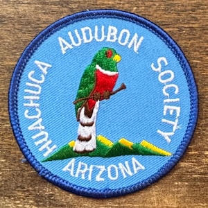 Puede incluir: Un parche redondo azul con el texto "Audubon Society" y "Huachuca Arizona" que rodea un pájaro colorido posado en una rama. El pájaro tiene plumas verdes, rojas y blancas, con un pico amarillo. El parche es bordado.