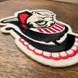 Pu&ograve; includere: Una toppa ricamata raffigurante un personaggio stilizzato con volto color crema, un cappello nero con un accento rosso e un ampio sorriso con denti a strisce rosse e bianche. Il design &egrave; delineato da filo bianco con un bordo smerlato rosso.
