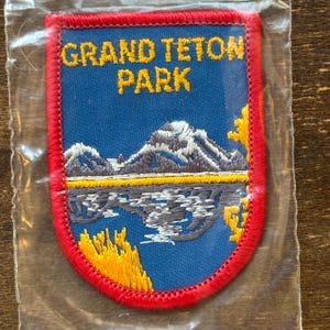 May include: Embroidered patch with the text "GRAND TETON PARK" in yellow on a blue background. The patch features a mountain range and a lake scene, with a red border. The patch is in a clear plastic bag.
