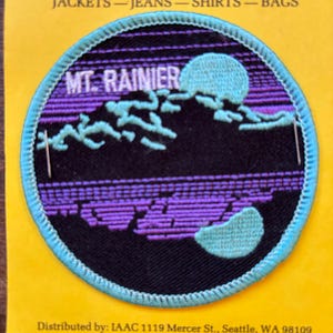 Pode incluir: Um remendo bordado azul e roxo com o texto "MT. RAINIER" e uma cena de montanha. O remendo tem uma borda azul.