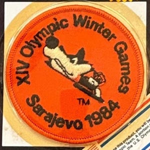 May include: A circular orange embroidered patch featuring the black text XIV Olympic Winter Games Sarajevo 1984. In the center, a white and black stylized wolf mascot graphic is depicted. The patch has a stitched border and a TM symbol.