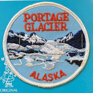 May include: Round embroidered patch featuring a mountain landscape with the words "Portage Glacier Alaska" in orange lettering on a light blue background. The patch has a white border and depicts snow-capped mountains and a body of water.