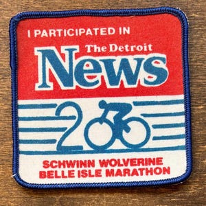 Puede incluir: Parche cuadrado con fondo rojo y blanco, borde azul y texto. El texto dice "I PARTICIPATED IN The Detroit News 2006 SCHWINN WOLVERINE BELLE ISLE MARATHON" con un gráfico de bicicleta azul.