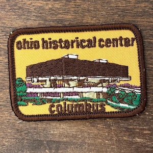 Pode incluir: Patch bordado com o Ohio Historical Center em Columbus. O patch tem um fundo amarelo, uma borda marrom e texto. O edifício é marrom com detalhes brancos e folhagem verde e roxa.