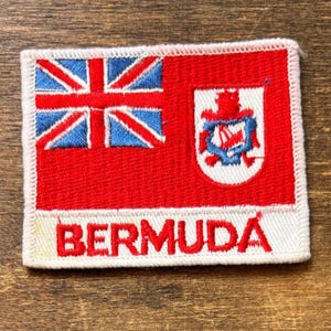 May include: A rectangular Bermuda flag patch with a white border. The flag features a Union Jack in the upper left corner, a red field, and the Bermuda coat of arms. The word "BERMUDA" is embroidered in white.