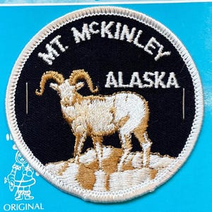 May include: Circular patch featuring a ram with large curved horns, standing on a rocky landscape. The patch is embroidered with the words "MT. MCKINLEY ALASKA" in white lettering against a black background, with a white border.